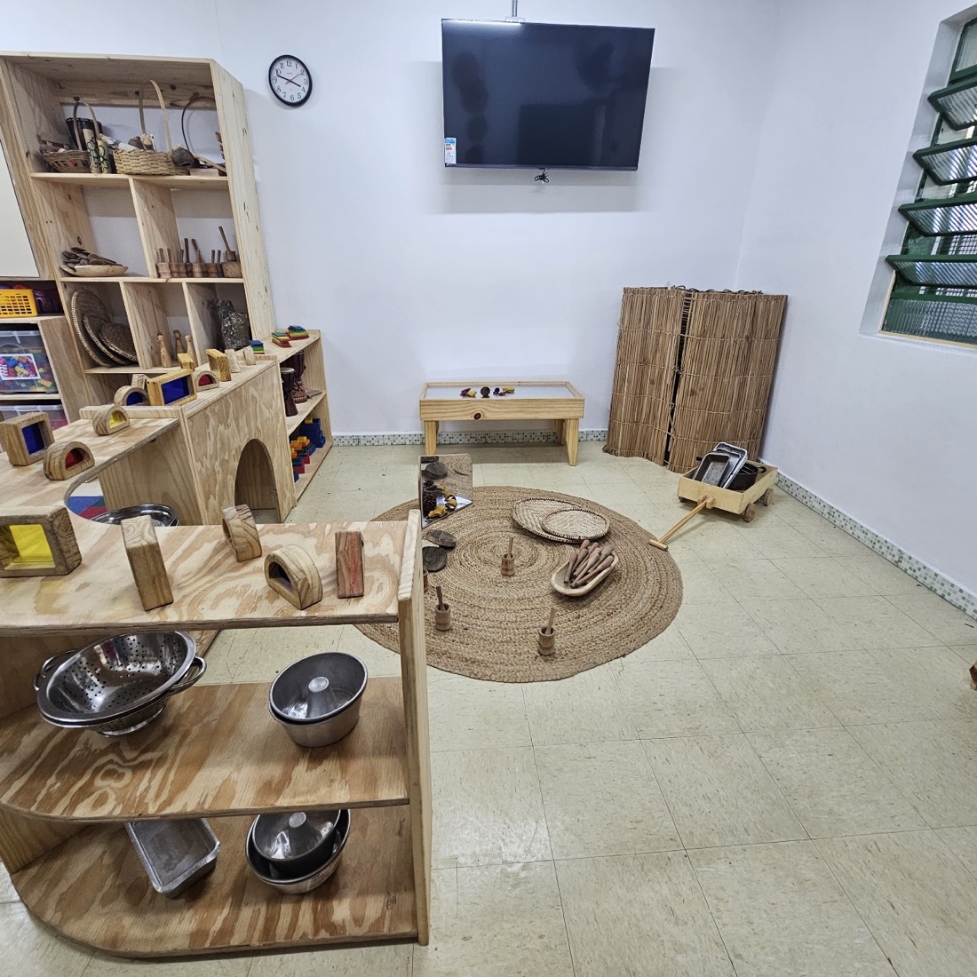 Cantinho de exploração com estante alta de nichos, bancada curva com utensílios de metal e tapete de juta com materiais soltos.