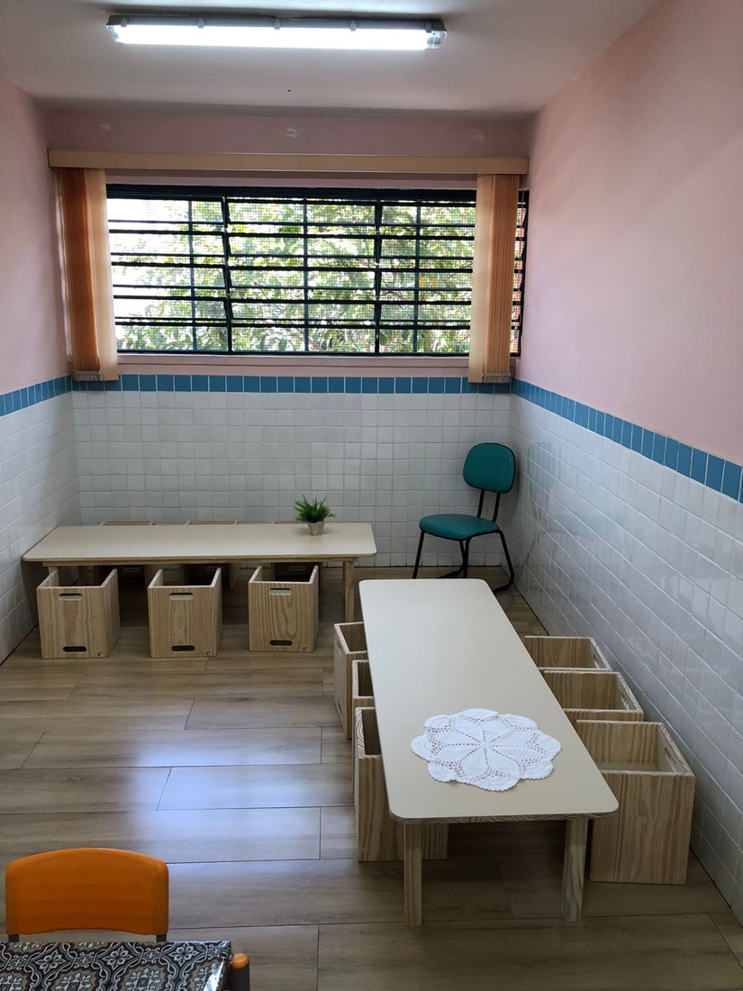 Sala com paredes rosa e azulejo branco, duas mesas coletivas em madeira e caixotes usados como assento e organização.
