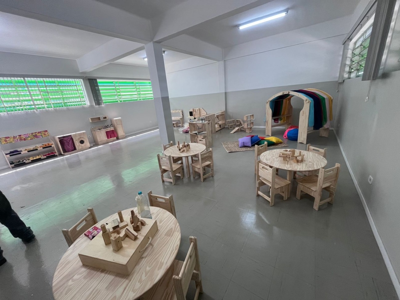 Sala ampla com mesas redondas e cadeiras infantis, cozinha de brinquedo, prateleiras baixas e cabana de madeira com tecidos coloridos.