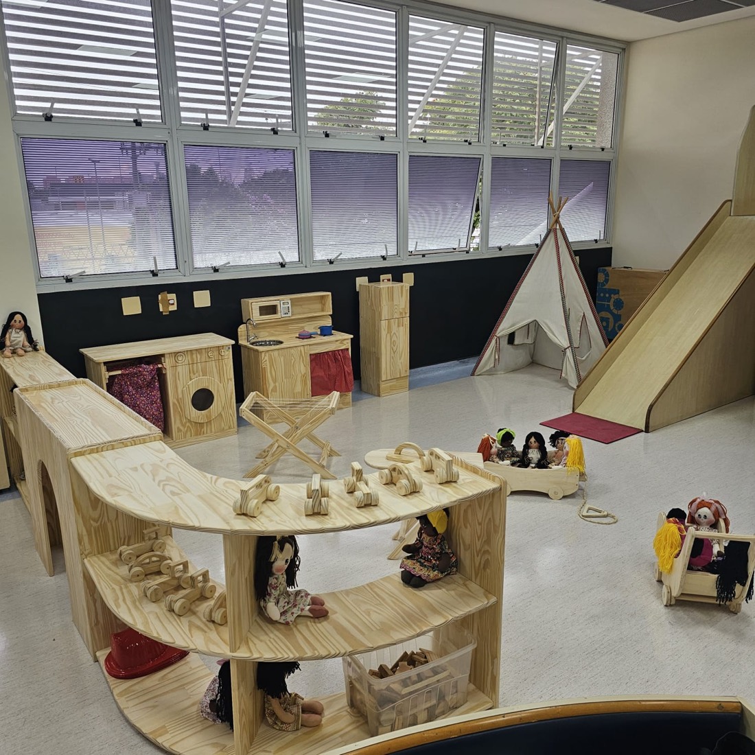 Brinquedoteca com prateleira curva cheia de carrinhos e bonecas, escorregador de madeira, tenda e cozinha de brinquedo.