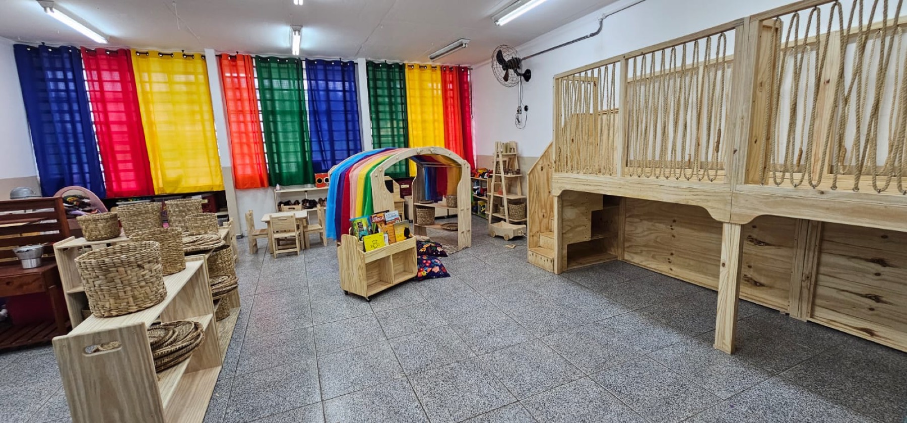 Sala de educação infantil com estrutura elevada em madeira com guarda-corpo de cordas, cabana arco-íris, prateleiras baixas com cestos e mesas infantis.