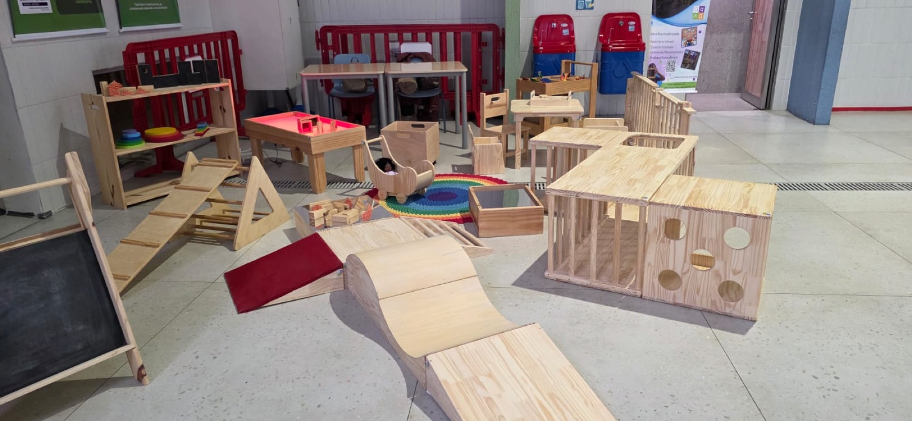 Conjunto de peças do Ateliê montadas lado a lado: rampa curva, triângulo de escalada, casinha de madeira, mesa de luz, lousa e carrinhos.