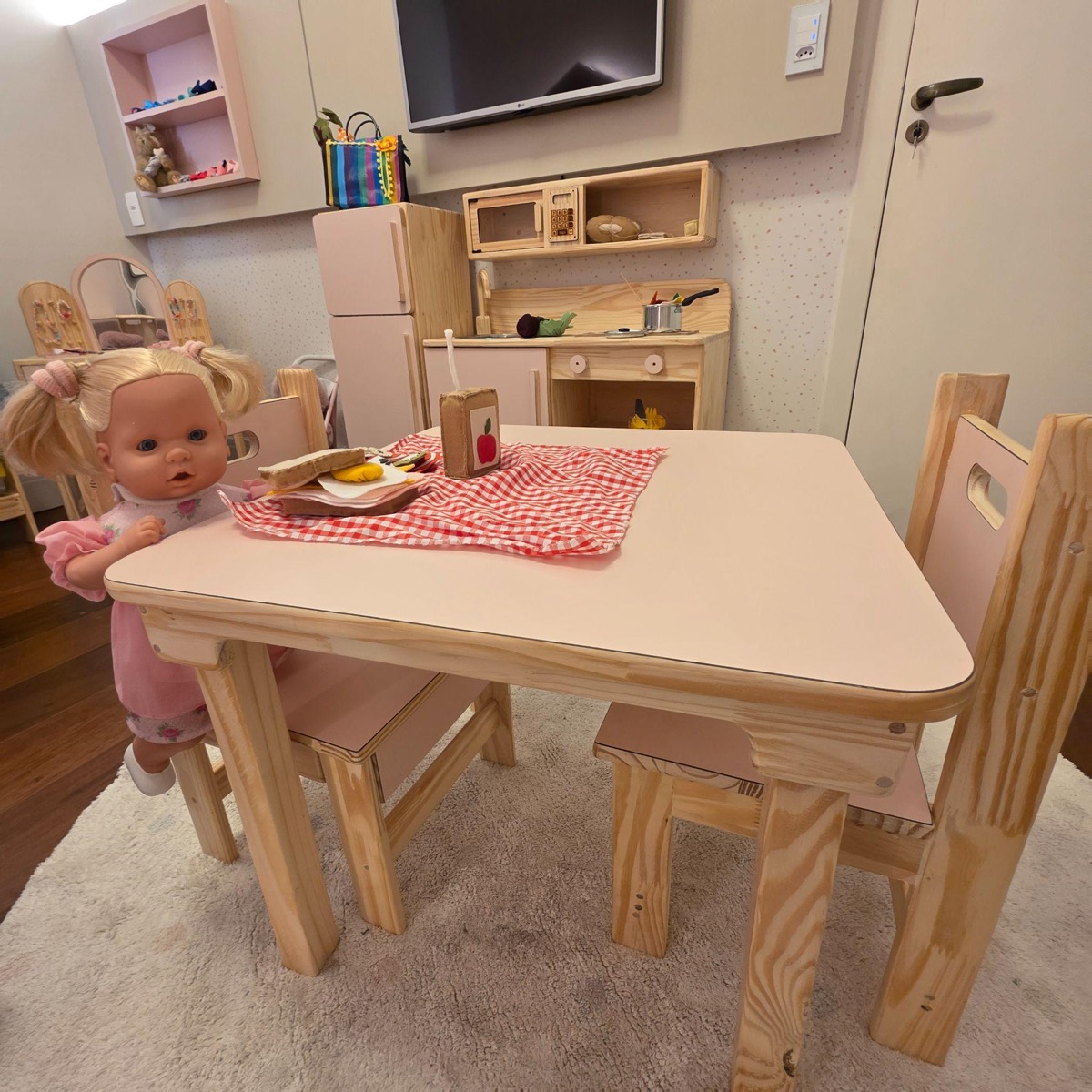 Mesa e cadeiras infantis de madeira com toalha xadrez e comidinhas de brinquedo, ao fundo geladeira e cozinha de madeira.