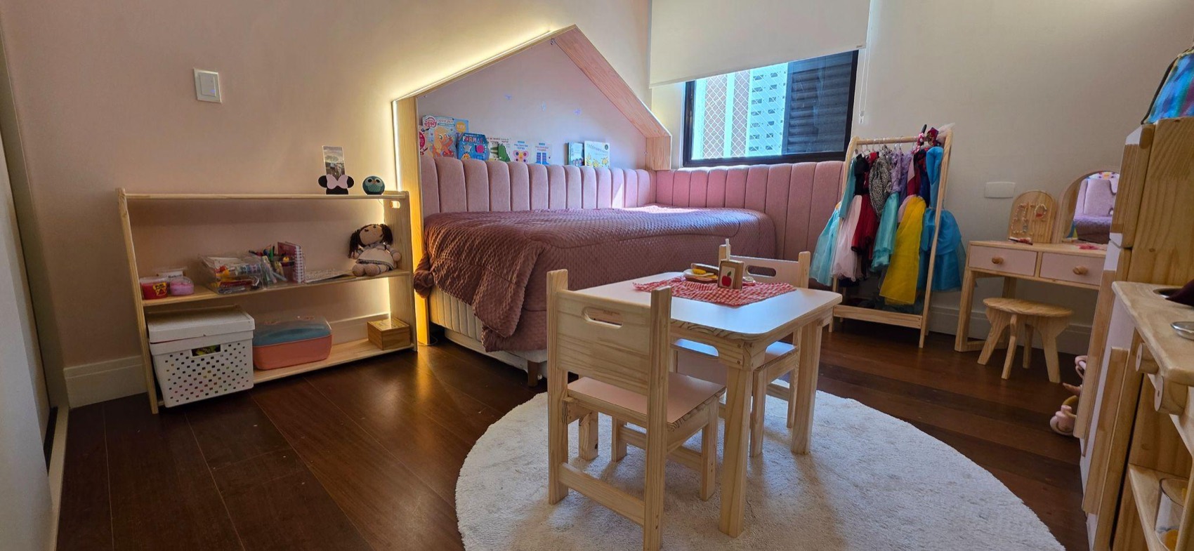 Quarto infantil montado com estante baixa de brinquedos, cama com cabeceira em formato de casinha, mesa e cadeiras infantis sobre tapete redondo e arara de fantasias.